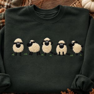 Op de afbeelding: Een donker bosgroene sweater met ronde hals voorzien van een borduursel van vijf pluizige, crèmekleurige schapen met zwarte gezichten en poten. De schapen staan in verschillende houdingen op de borst, afgebeeld op kleine groene graspolletjes.