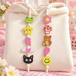 Puede incluir: Una sudadera con capucha de color crema con cordones blancos gruesos adornados con varios parches bordados. El cordón izquierdo muestra una rana verde, una estrella amarilla, medio corazón rosa, un corazón alado y un gato negro. El cordón derecho muestra una flor rosa, un hongo rojo, un oso marrón, medio corazón rosa y una cara sonriente amarilla.