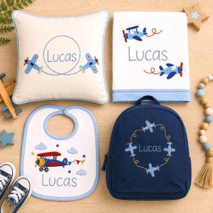May include: A set of personalized baby items including a cream square pillow, a white waffle-weave blanket, a white bib with blue trim, and a navy blue backpack. Each item features embroidered blue airplanes and the name Lucas in blue thread.
