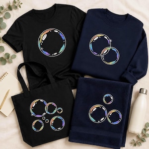 Pode incluir: Uma t-shirt preta com um gráfico de uma única bolha, uma camisola azul-marinho com dois gráficos de bolhas sobrepostas, um saco de pano preto com cinco gráficos de bolhas e uma toalha azul-marinho com três gráficos de bolhas iridescentes.