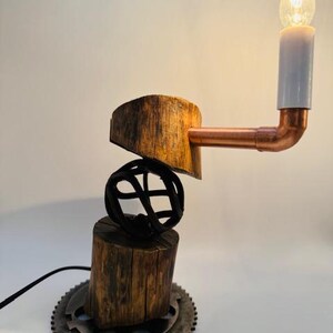 May include: A rustic table lamp crafted from reclaimed wood, metal, and copper pipe. The lamp features a unique design with a round metal base, a wooden cylinder, and a copper pipe arm with a light bulb. The lamp is lit and casting a warm glow.