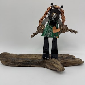 May include: A quirky robot sculpture made from recycled components. The robot has a circuit board body, key arms, and copper wire hair. It is mounted on a piece of driftwood, displaying a unique mix of industrial and natural materials.