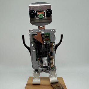 May include: A handcrafted robot sculpture made from repurposed metal and electronic components. The robot features a silver head, black eyes, and a green center. Its body is constructed from a circuit board, with arms and legs attached to a wooden base.