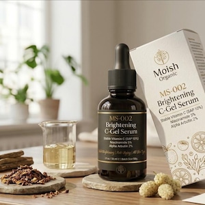 May include: A dark amber glass dropper bottle of Moish Organic MS-002 Brightening C-Gel Serum with a black cap, accompanied by its white rectangular box. The label and box list Stable Vitamin C (SAP 10%), Niacinamide 5%, and Alpha Arbutin 2%.