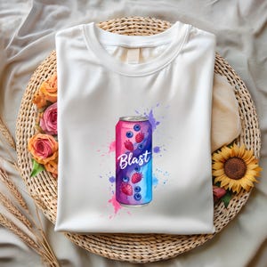 May include: White t-shirt with a vibrant graphic of a drink can. The design features the word "Blast" and illustrations of blueberries and strawberries, set against pink and blue watercolour splashes.