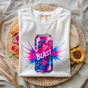 May include: White t-shirt with a vibrant graphic of a soda can. The can design features the word "BLAST" and illustrations of blueberries, strawberries, and raspberries, with a pink and blue splash effect.
