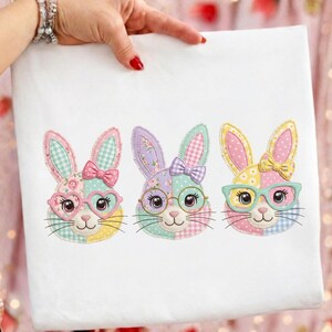 May include: White fabric featuring three embroidered bunny faces. Each bunny has a unique patchwork design, bow, and glasses. The bunnies are in pastel colors, including pink, yellow, and purple, with floral and gingham patterns.