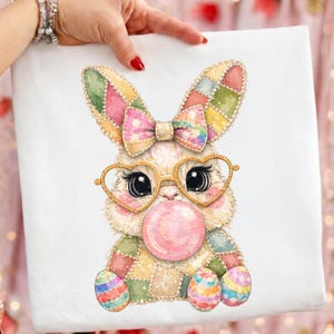 Pode incluir: Tela branca com um coelho de desenho animado com óculos em forma de coração soprando uma bolha de chiclete. O coelho tem um design de patchwork com um laço e ovos de Páscoa coloridos. A imagem é segurada por uma pessoa.