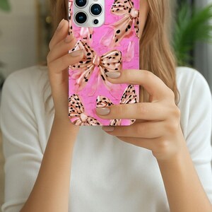 May include: A pink phone case featuring a pattern of bows with a leopard print design. The bows have a metallic gold color and are set against a textured pink background. The case has a glossy finish.