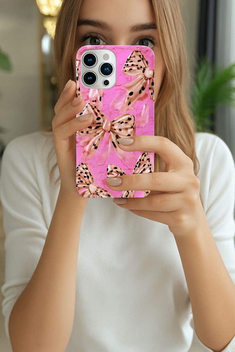 May include: A pink phone case featuring a pattern of bows with a leopard print design. The bows have a metallic gold color and are set against a textured pink background. The case has a glossy finish.