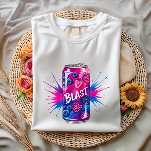 May include: White t-shirt with a vibrant graphic of a soda can, the word "BLAST" is printed on the can. The can design includes blueberries, strawberries, and raspberries, with a pink and blue splash effect. The shirt is on a woven basket.