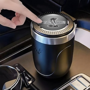 May include: Black and silver car ashtray with the text "SHELBY GT 500 SUPER SNAKE" and a cobra logo. The ashtray has a circular top with a silver rim and a black body. A finger is pressing the top of the ashtray.