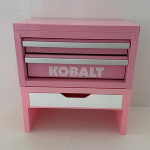 May include: A pink metal tool chest with two drawers featuring silver handles and the white text KOBALT printed on the front. Below the metal chest is a separate pink stand with a single white drawer and an open storage space at the bottom.
