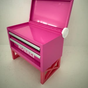 May include: A bright pink metal toolbox with a hinged lid and two drawers. The drawers have silver trim and the word "KOBALT" is visible on the front. The toolbox sits on a matching pink and red base.