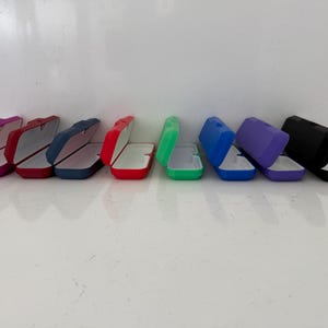 May include: A collection of open eyeglass cases in various colors, including pink, red, navy, green, blue, purple, and black. Each case is rectangular with a hinged lid and a white interior, suitable for storing eyewear.