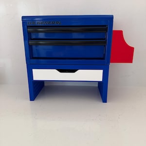 May include: A blue US General tool chest with three black-handled drawers and a white drawer. The chest has a red side panel and is on a white surface. The text "US GENERAL" is visible on the top.