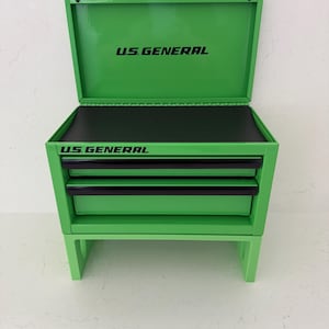 May include: A bright green U.S. GENERAL tool chest with two drawers and a top compartment. The open top reveals a black interior. The words "U.S. GENERAL" are printed in black on the top and front of the chest.