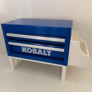 May include: A blue and silver tool chest with the word "KOBALT" in white lettering. The tool chest has two drawers and a white side compartment. The chest is supported by white legs.
