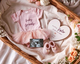 May include: A pink baby bodysuit with the name "Baby Brooks" printed on it, paired with a tulle skirt, baby shoes, and a heart-shaped sign that reads "A Little Sister is on the way! June 2026." The items are arranged in a wicker basket with floral accents.
