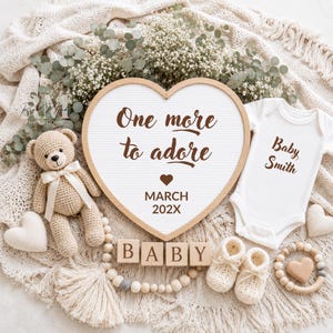 Może przedstawiać: Tablica w kształcie serca z napisem One more to adore MARCH 202X, białe body dziecięce z nadrukiem Baby Smith, szydełkowany pluszowy miś z kremową wstążką, cztery drewniane klocki z literami BABY oraz kremowe buciki niemowlęce.