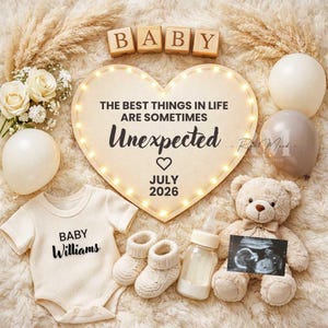 May include: A baby announcement with a cream-colored onesie, booties, and a teddy bear. A heart-shaped sign with the text "THE BEST THINGS IN LIFE ARE SOMETIMES Unexpected" and "July 2026" is illuminated with lights. Wooden blocks spell "BABY".