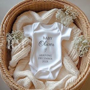 May include: White baby onesie with the text "BABY Oliver" and a heart symbol, announcing an arrival in December 202X. The onesie is placed in a woven basket with a cream-colored blanket and small white flowers.