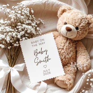 May include: A soft, plush teddy bear next to a white card with the text "Baby on the Way" and "Baby Smith July 2026." A bouquet of white flowers with a white ribbon is also in the image. The overall theme is a baby announcement.