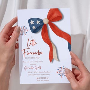 Könnte beinhalten: Eine Babyparty-Einladung mit patriotischem Thema. Die Karte zeigt eine Aquarellschleife in Rot, Weiß und Blau mit dem Text "A Little Firecracker Is On The Way". Zusätzlicher Text enthält den Namen der Geehrten, Datum, Uhrzeit und Ort.
