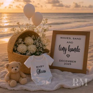 以下が含まれることがあります： ビーチでのベビー発表。 「Baby Olivia」とプリントされた白いベビー服が、テディベアと「Waves, Sand and tiny hands December 2025」と書かれた額入りの看板の隣にあります。 2つの白い風船と花が編まれたバスケットに入っています。