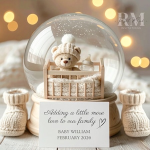 May include: A snow globe with a teddy bear in a miniature crib, a baby bottle, and a sign that reads "Adding a little more love to our family" with the name "Baby William" and the date "February 2026". Two knitted baby booties are on either side.