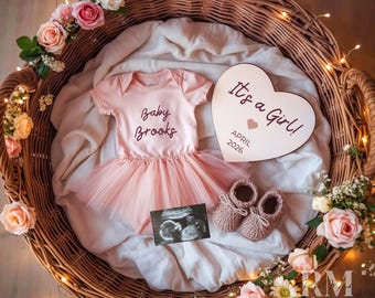 May include: A pink baby onesie and tutu skirt set with the text "Baby Brooks" inside a wicker basket. A wooden heart-shaped sign reads "It's a Girl!" and "April 2026". Pink baby booties and ultrasound image complete the scene.