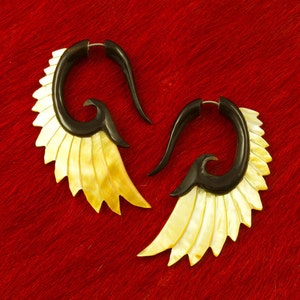 May include: A pair of unique earrings with a wing design. The earrings feature a black, curved top and a fan of cream-colored, shell-like segments. The earrings are displayed on a red background.