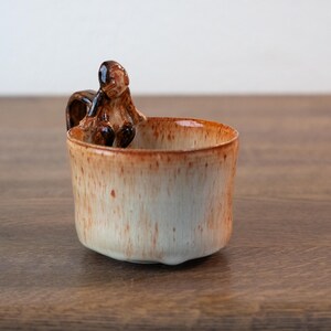 Pode incluir: Uma caneca de cerâmica com uma figura de esquilo na borda. A caneca tem um exterior salpicado em branco sujo e castanho. O esquilo é detalhado com detalhes em castanho escuro.