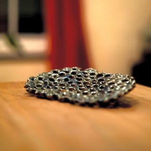May include: A small, irregularly shaped, metallic dish with a textured surface. The dish is silver-toned with numerous small, dark holes throughout. The object is set on a wooden surface, with a red curtain in the background.