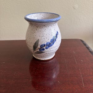 May include: A small, bulbous ceramic cup with a speckled white glaze and a blue rim. The exterior features a hand-painted design of a blue floral sprig with green leaves. The cup has a flared opening and a narrow base, crafted from stoneware.