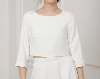 Puede incluir: Conjunto blanco de dos piezas con top corto de manga tres cuartos y falda a juego. El top tiene cuello barco. El conjunto está confeccionado en tejido liso y opaco. La modelo sonríe.