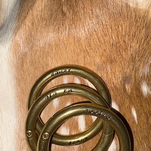 May include: Three brass key rings with engraved text, overlapping slightly. The rings have the words "HOWDY", "GIG EM", and "AGGIES" etched into them. The background features a brown and white cowhide pattern. The rings are made of brass.