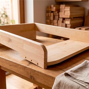 Peut inclure: Un plateau à langer en bois clair avec une base rectangulaire et des panneaux latéraux surélevés. Le plateau présente une finition en grain de bois naturel lisse, des bords arrondis et une construction robuste conçue pour reposer sur une surface plane.