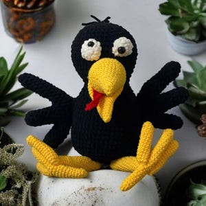 Puede incluir: Un cuervo de crochet negro con pico y patas amarillas. El cuervo tiene grandes ojos blancos y una lengua roja que sobresale. El cuervo está sentado sobre una superficie blanca.