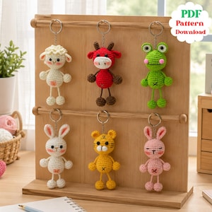 May include: Six crocheted animal keychains hanging on a wooden display stand. The figures include a white sheep, a red cow, a green frog, a white rabbit, a yellow tiger, and a pink rabbit. Each keychain features a metal ring and a PDF Pattern Download label.