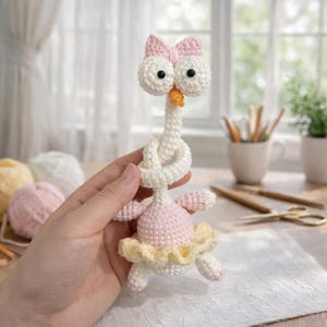 May include: A handmade crochet swan doll with a pink tutu and bow. The swan has large, round eyes and an orange beak. The doll is made of white, pink, and yellow yarn. The swan's neck is tied in a knot.