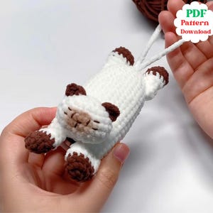 Op de afbeelding: Een handgemaakt gehaakt kattenspeeltje in wit en bruin. De kat ligt met uitgestrekte poten. De oren, poten en het gezicht van de kat hebben bruine accenten. De afbeelding bevat een witte, wolk-vormige afbeelding met de tekst "PDF Pattern Download".