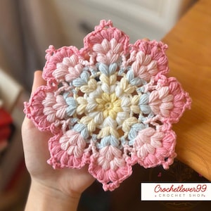 May include: A handmade, multi-colored crochet coaster in a floral design. The coaster features a central yellow flower surrounded by petals in shades of pink, white, and blue. The edges are a darker pink. The text "Crochetlover99 + Crochet Shop" is in the bottom right corner.
