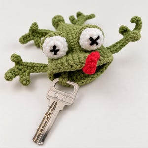 Puede incluir: Una funda para llave de ganchillo verde con forma de rana, con dos grandes ojos blancos con pupilas en forma de X negra y una pequeña lengua roja. La rana tiene cuatro extremidades extendidas y sujeta una llave de metal plateado con las letras ON.