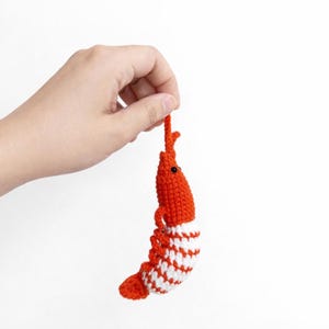 May include: A handmade, crocheted shrimp toy in bright orange and white. The shrimp has black eyes and is held by a hand against a white background. The shrimp's body is orange with a white and orange striped tail.