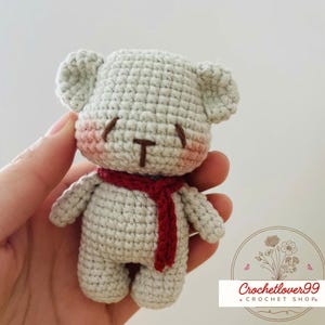 May include: A small, crocheted teddy bear in light gray with a red scarf. The bear has rosy cheeks and a brown nose and mouth. The image includes the text "Crochetlover99 + Crochet Shop."