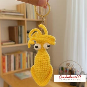 May include: A handmade yellow crochet keychain featuring a whimsical creature with large, googly eyes. The keychain has a gold-colored ring and chain. The background includes a bookshelf and the text "Crochetlover99 + Crochet Shop."