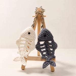 May include: Two crochet fish skeleton keychains, one in off-white and one in dark gray, each featuring a small black bead eye. The fish are attached to a silver-toned star-shaped metal keyring with a lobster clasp, showcasing intricate yarn work.