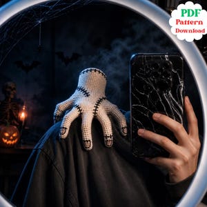 May include: A crocheted hand figurine in off-white yarn with black stitched details resembling scars and fingernails. The hand is positioned on a dark fabric surface. A digital file label in the corner reads PDF Pattern Download.