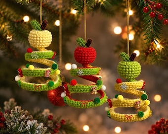 May include: Three hanging crochet ornaments shaped like spiral Christmas trees topped with crocheted apples. The trees are yellow and green, decorated with small colorful pom-poms, metallic charms, and snowflake accents, suspended by thin gold strings.